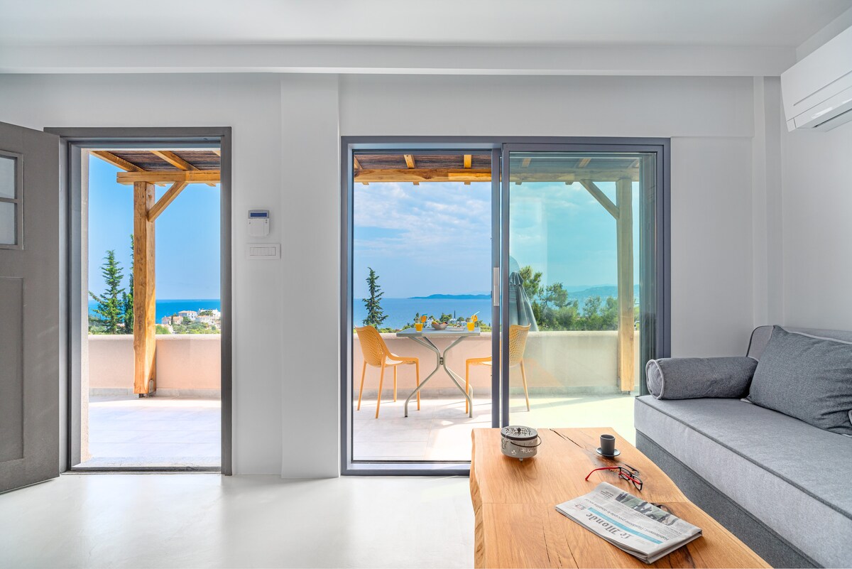 A spacious living area features large glass doors that open to a balcony, offering views of the sea. A gray sofa is positioned near a wooden coffee table. The light-filled space is complemented by an airy atmosphere and casual dining set on the terrace.