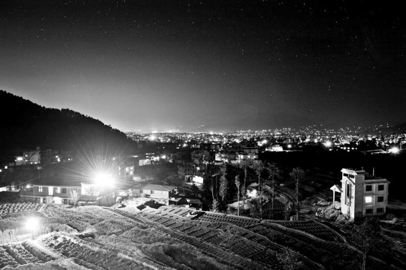 A nighttime view captures the illuminated landscape of the capital city, with twinkling lights stretching across the horizon. The surrounding hills are silhouetted against a starry sky, and the foreground showcases terraced fields and sparse structures, creating a tranquil atmosphere.