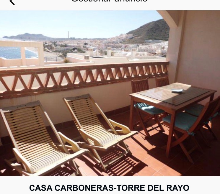 A balcony is presented with two wooden lounge chairs and a rectangular dining table, set against a backdrop of coastal views. The floor is tiled, and the railing features wooden slats, enhancing the outdoor space's inviting aspect.