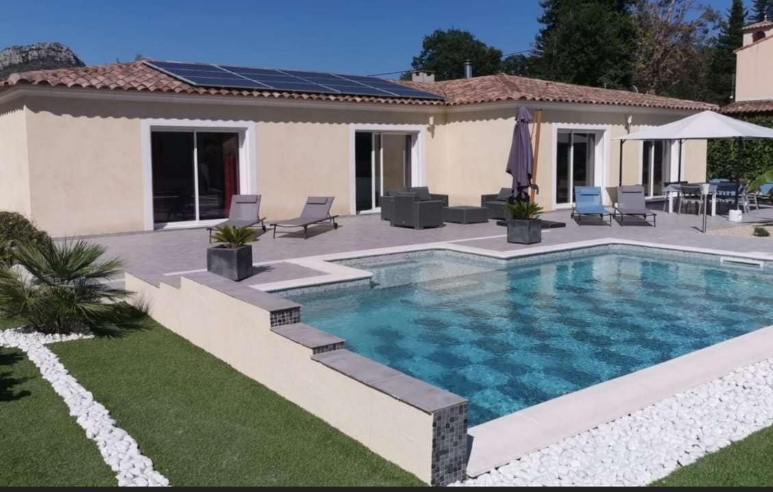 A contemporary villa is featured with an inviting private pool, surrounded by a patio area furnished with tables and chairs. The exterior is complemented by trimmed greenery and decorative stones, and sun loungers are positioned near the pool under a large umbrella.