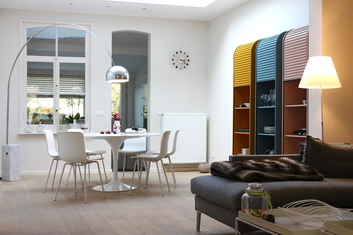 A bright dining area is featured, showcasing a round table surrounded by sleek white chairs. Colorful cabinets are accentuated by a contemporary floor lamp and a unique arc-shaped light fixture. Large windows allow natural light to fill the space, creating a warm and welcoming ambiance.