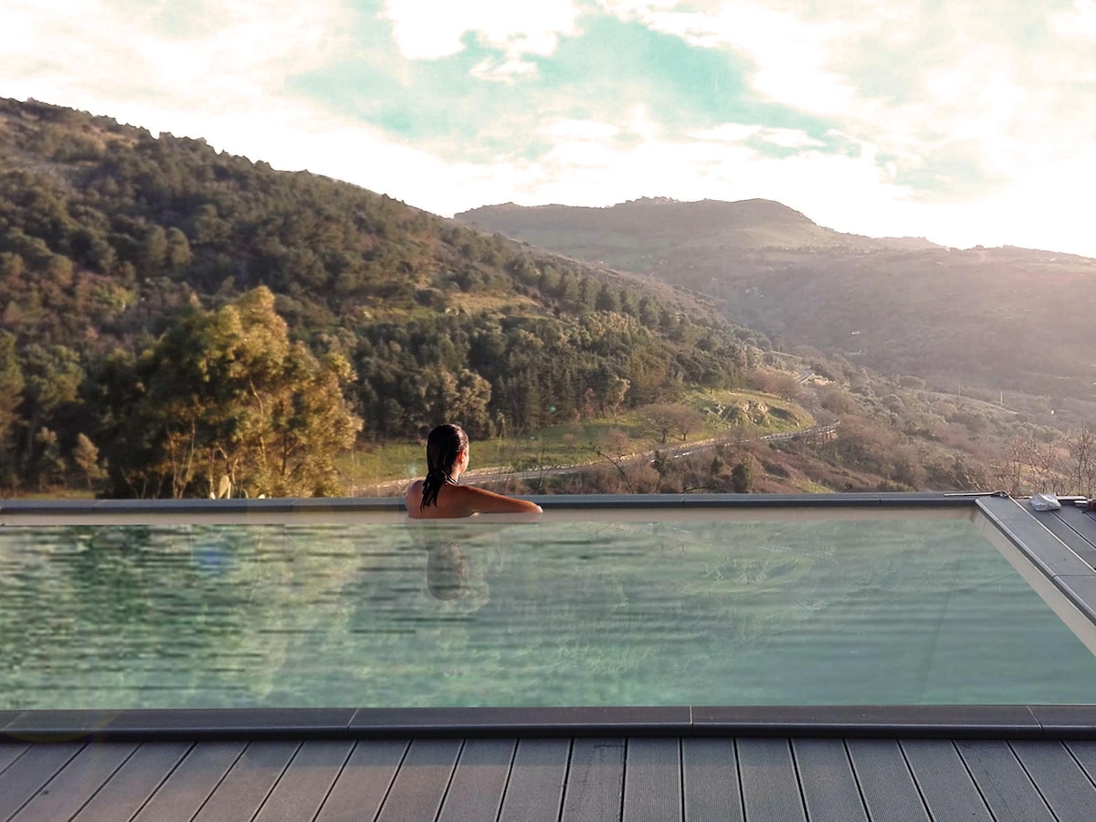 An infinity pool stretches across a scenic landscape, with mountains and valleys visible in the background. The tranquil water reflects the sky, creating a seamless view of nature. A person relaxes at the edge, enhancing the peaceful ambiance of the setting.