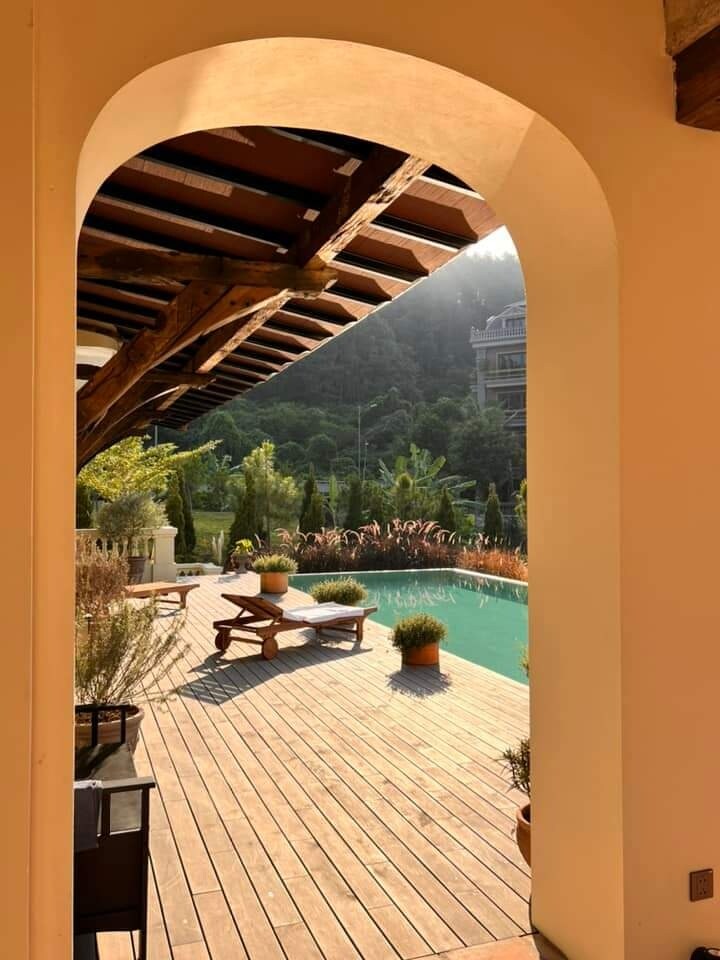 An outdoor area is framed by an archway, leading to a wooden deck. Plush seating and potted plants are arranged beside a tranquil pool, surrounded by lush greenery and distant hills. Soft sunlight filters through, creating a serene ambiance for relaxation.