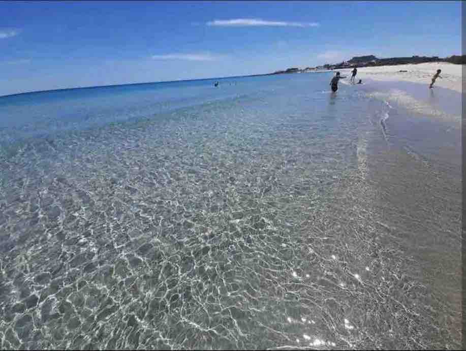 A clear and calm shoreline is visible, with gentle waves lapping against the sand. The water exhibits a bright, transparent hue, reflecting the sky. A few people can be seen enjoying the beach, surrounded by an expansive stretch of soft sand.