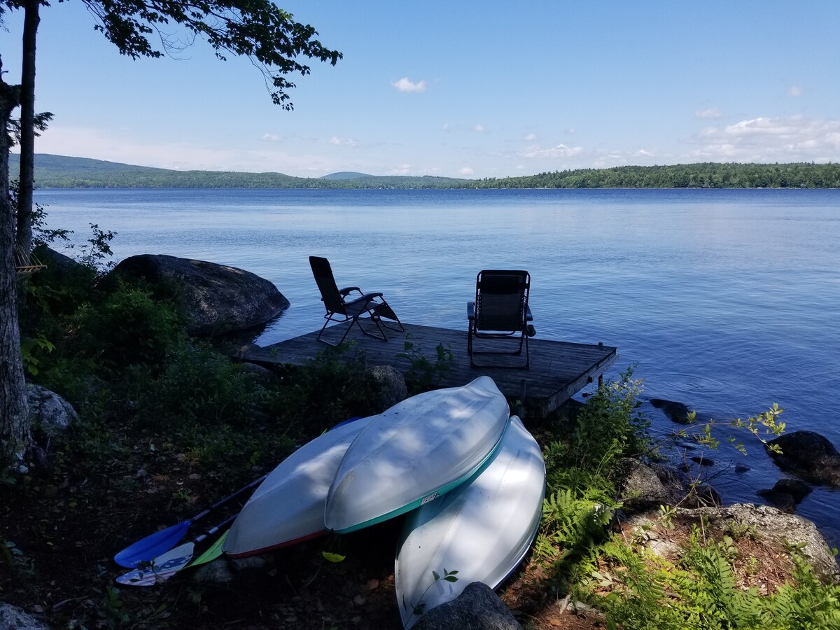A lakeside dock is depicted, with two lounge chairs positioned for serene views of the water. Several kayaks and paddleboards rest on the shore, framed by lush greenery and rocky terrain. The scene captures a peaceful moment under a bright blue sky.