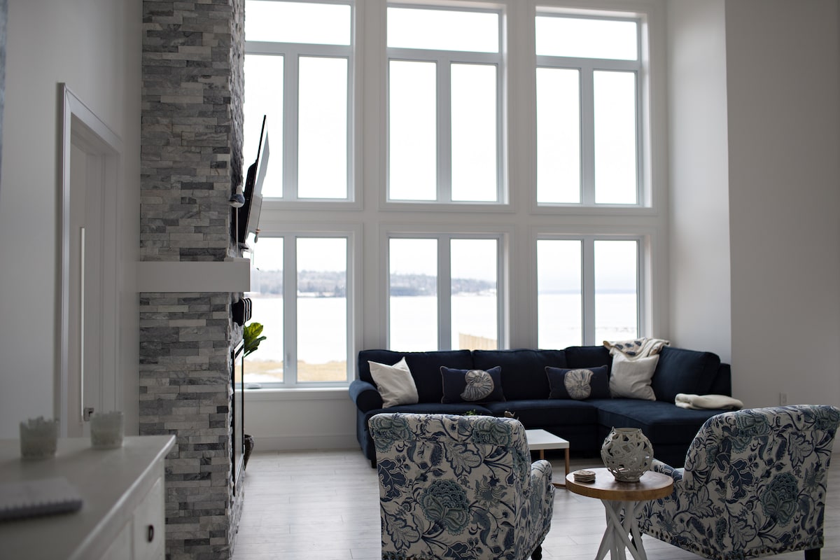 A light-filled living area features large windows that provide views of St. Margaret's Bay. Comfortable seating includes a deep blue sectional sofa adorned with patterned cushions, while two armchairs with floral patterns invite relaxation. A stone-accented fireplace adds an element of warmth to the space.