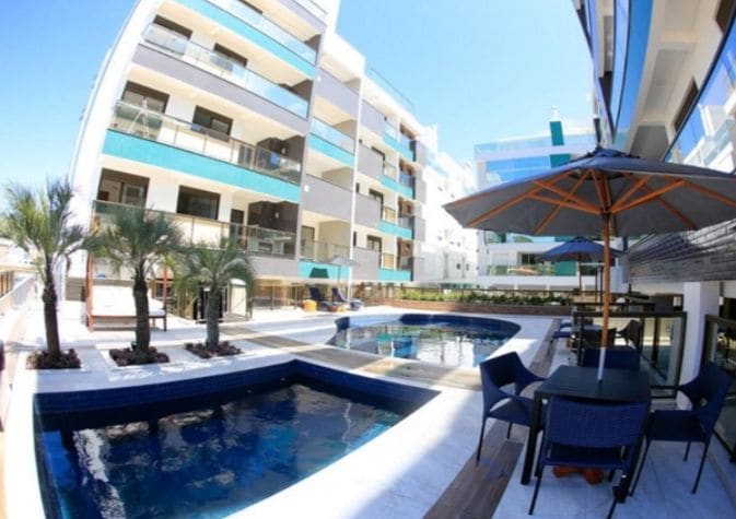 An outdoor area is depicted, featuring spacious swimming pools surrounded by palm trees. Lounge chairs and a shaded outdoor dining space are visible, offering a welcoming environment for relaxation. The modern architecture of the surrounding buildings enhances the overall appeal of the area.