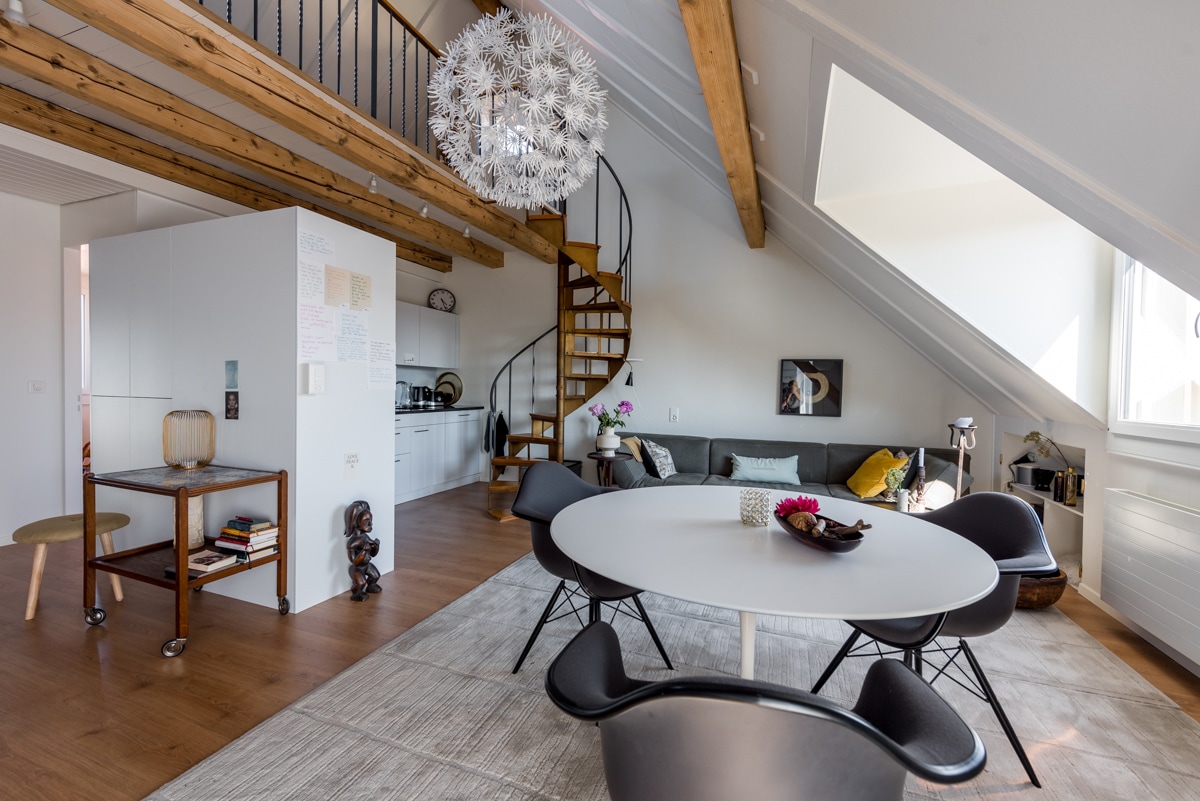 A spacious living area is highlighted by wooden beams and a spiral staircase. A round dining table is surrounded by black chairs, while a modern sofa provides a cozy seating option. Natural light floods the space through large windows, and decorative elements add a stylish touch.