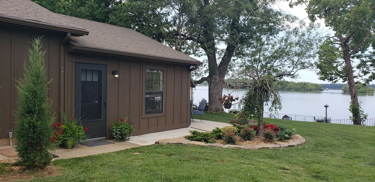 The exterior of the rental property is shown, featuring a brown wood-paneled facade. A well-maintained lawn surrounds the building, with vibrant flowerbeds adding color. A patio area with seating is visible by the lakeside, offering views of the water and trees.