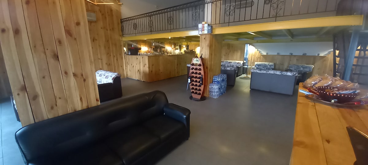 An open communal space features wooden paneling and grey tiled flooring. Comfortable sofas are arranged along the walls, with a display of wine bottles placed at the center. Soft lighting creates a relaxed environment, while various seating areas invite social interaction.