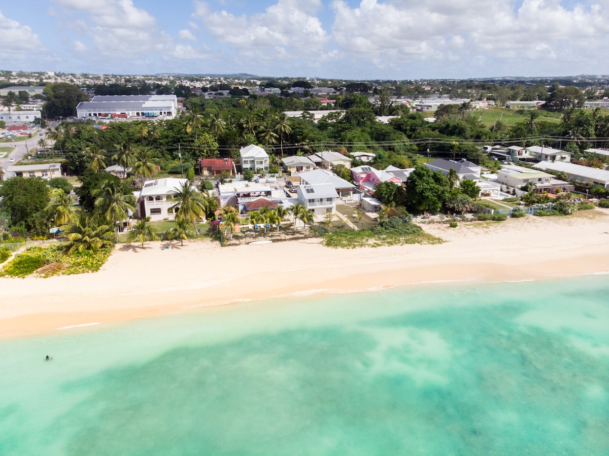 An aerial view showcases a sandy beach lined with palm trees and residential buildings. The turquoise water gently merges with the shore, while the lush greenery extends inland. The scene captures a tranquil coastal atmosphere, with homes nestled within the tropical surroundings.