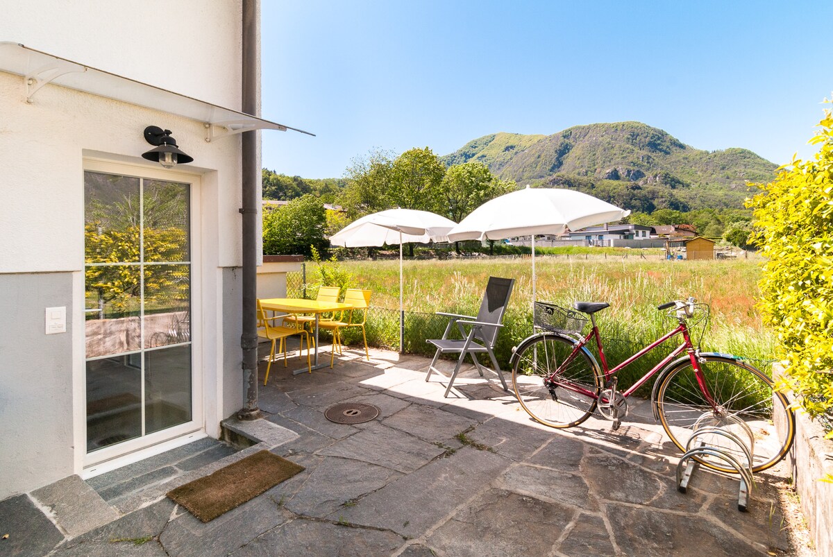 An outdoor patio area is presented, featuring a round yellow table and four matching chairs under two umbrellas. A red bicycle is positioned nearby, adjacent to a gray lounge chair, set against a backdrop of green fields and distant mountains under a clear blue sky.