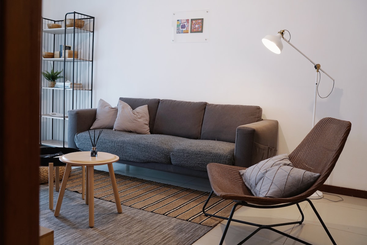 The living area features a modern grey sofa complemented by beige cushions. A light wooden coffee table sits in front, with a minimalist rug beneath. A sleek floor lamp provides illumination, and a decorative shelving unit displays plants and curated items.