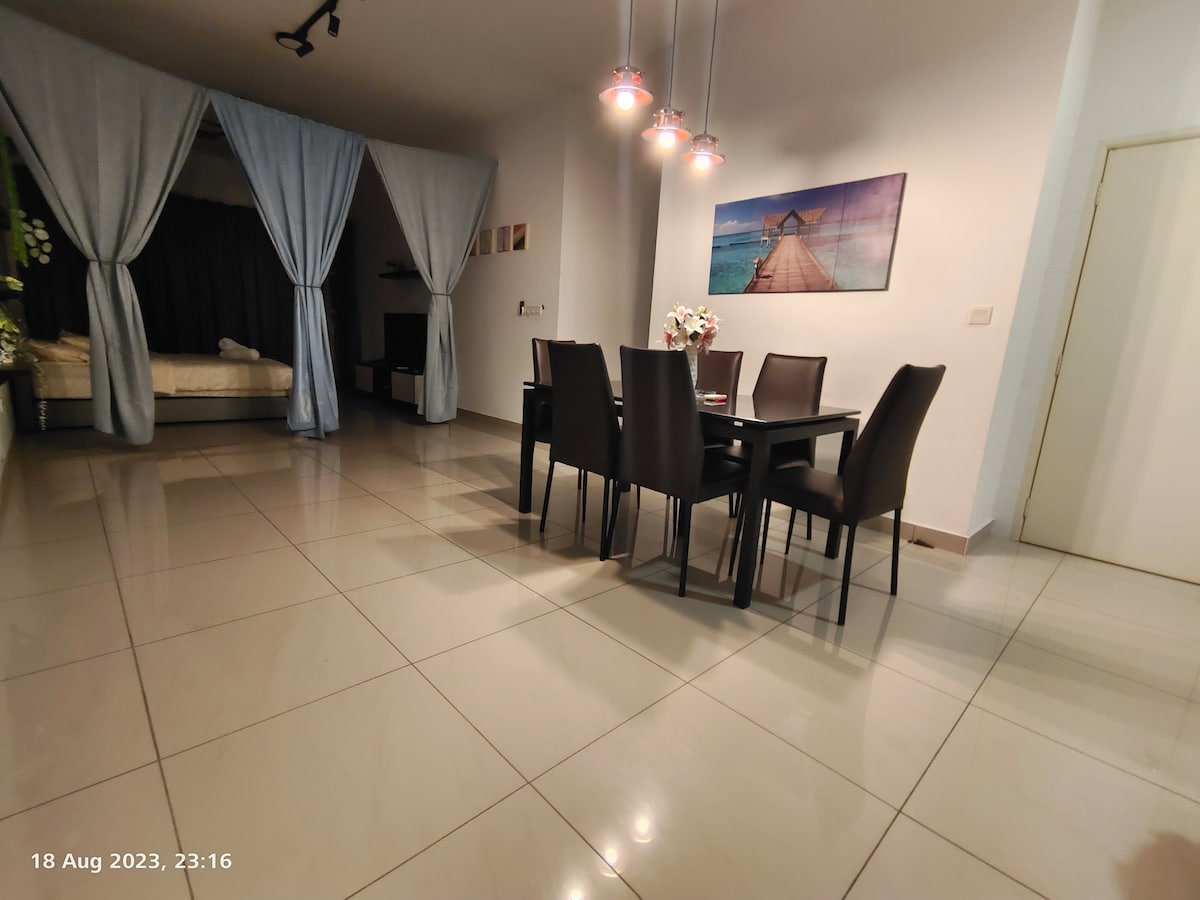 A dining area is showcased with a dark table surrounded by six chairs. Soft, blue drapes frame the space, while a photo depicting a serene waterfront scene is mounted on the wall. The floor reflects the overhead lighting, creating a bright atmosphere.
