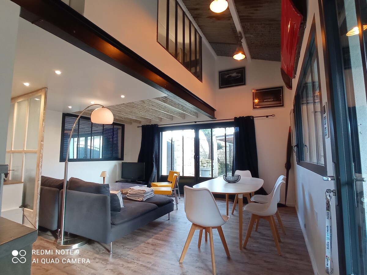 The spacious living area features modern furnishings, including a gray sofa, white dining table, and stylish chairs. Large glass doors open to a patio, allowing natural light to fill the room. Minimalist décor adds to the contemporary feel, while a wall-mounted TV is visible.