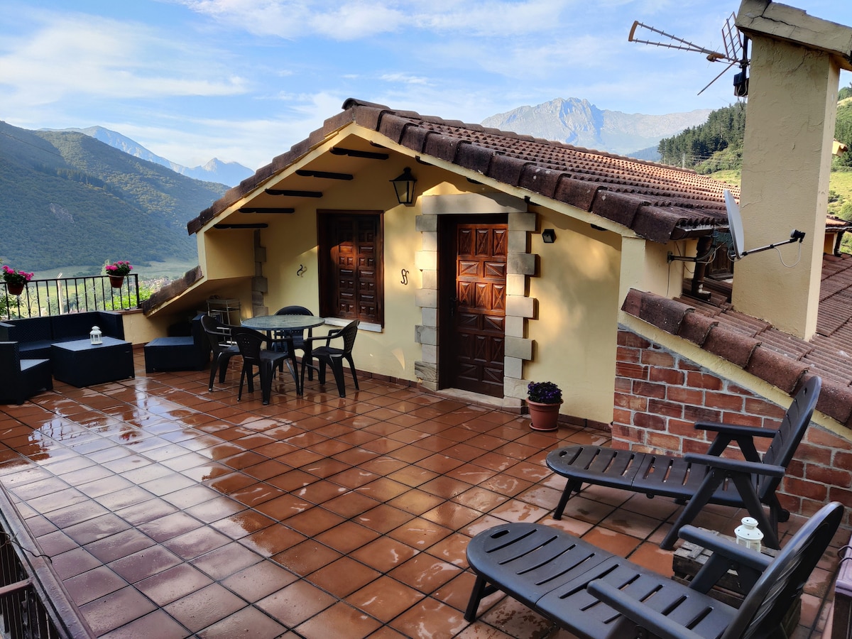 An expansive terrace is presented, featuring a tiled floor and surrounded by scenic mountain views. Outdoor seating options include a dining table with chairs and lounge chairs. The entrance to the house is visible, framed by wooden doors and decorative planters.