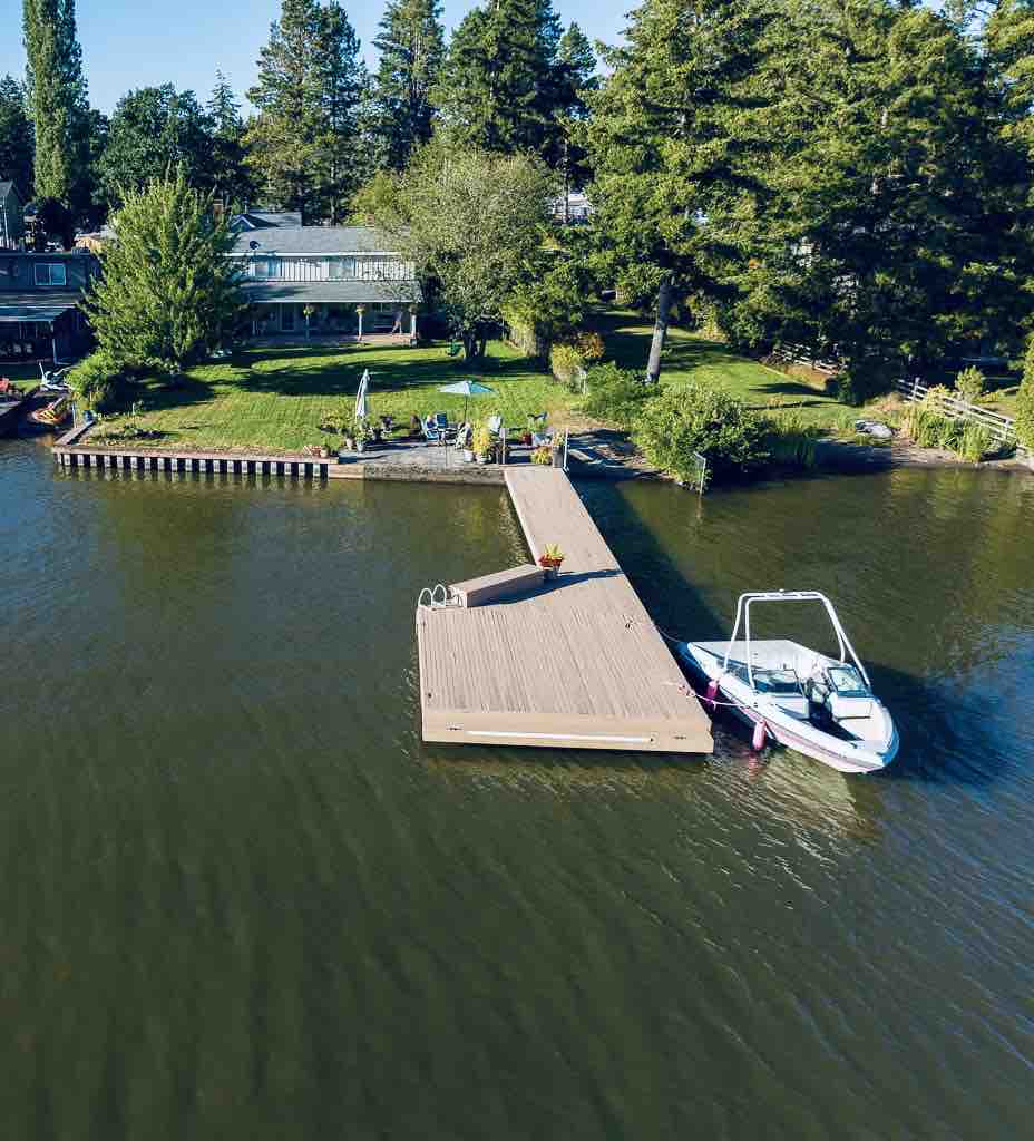 An aerial view captures a spacious private dock extending into the calm waters, with a boat moored at the end. Lush greenery surrounds the area, providing a natural setting, while picnic seating is visible on the lawn, offering a serene lakeside escape.