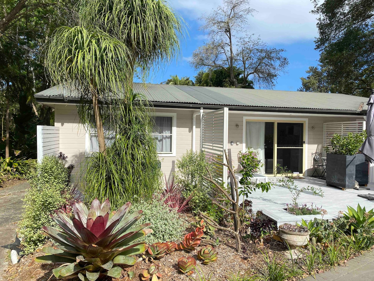 A private apartment is showcased with a neutral exterior and a sloped roof. Lush greenery and decorative plants frame the entry, which features large glass sliding doors leading to a spacious deck. The surrounding garden adds to the inviting ambiance.