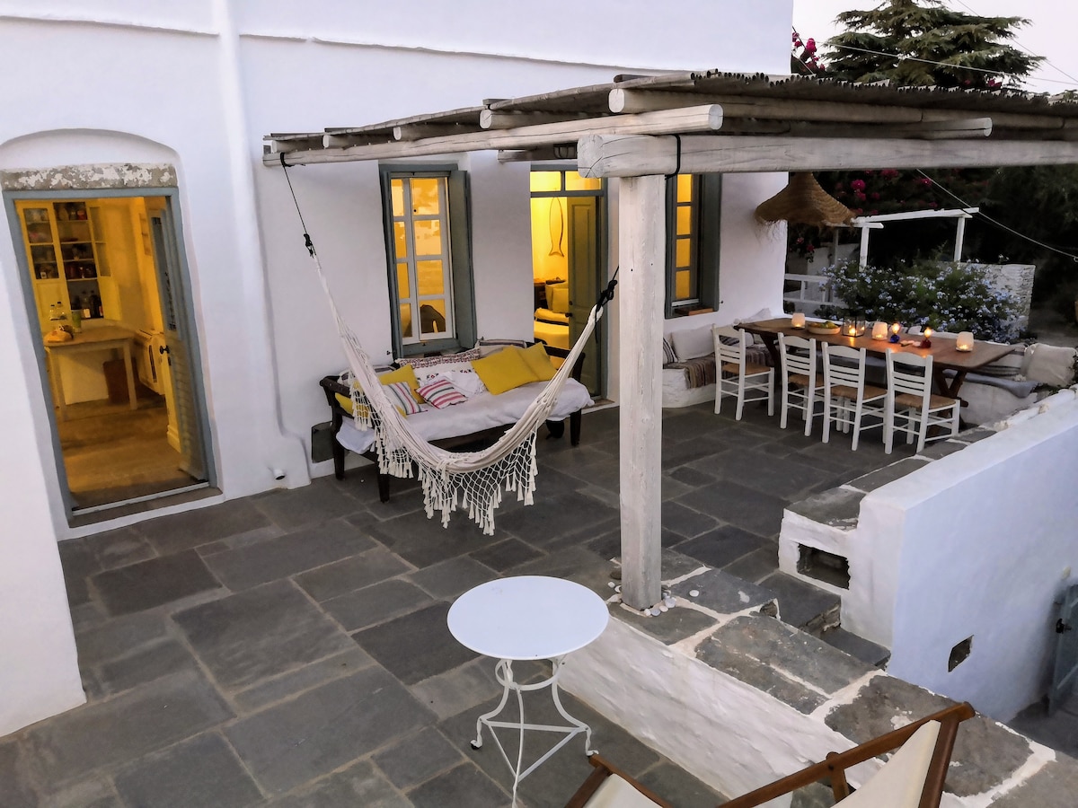 An outdoor terrace features stone flooring and a wooden pergola providing shade. A hammock is positioned nearby, while a dining table with multiple chairs is visible, set for an evening gathering. Soft lighting emanates from interior windows, enhancing the inviting atmosphere.