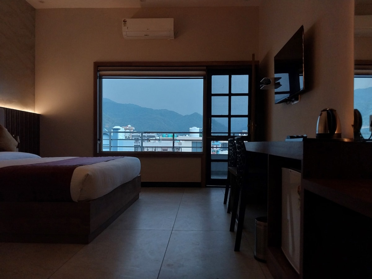 A well-lit bedroom features a spacious bed with neutral-toned bedding, along with a sleek workspace. A large window showcases a view of the mountains, complemented by dark wood accents and modern furnishings. An air conditioner and a flat-screen TV are visible on the wall.