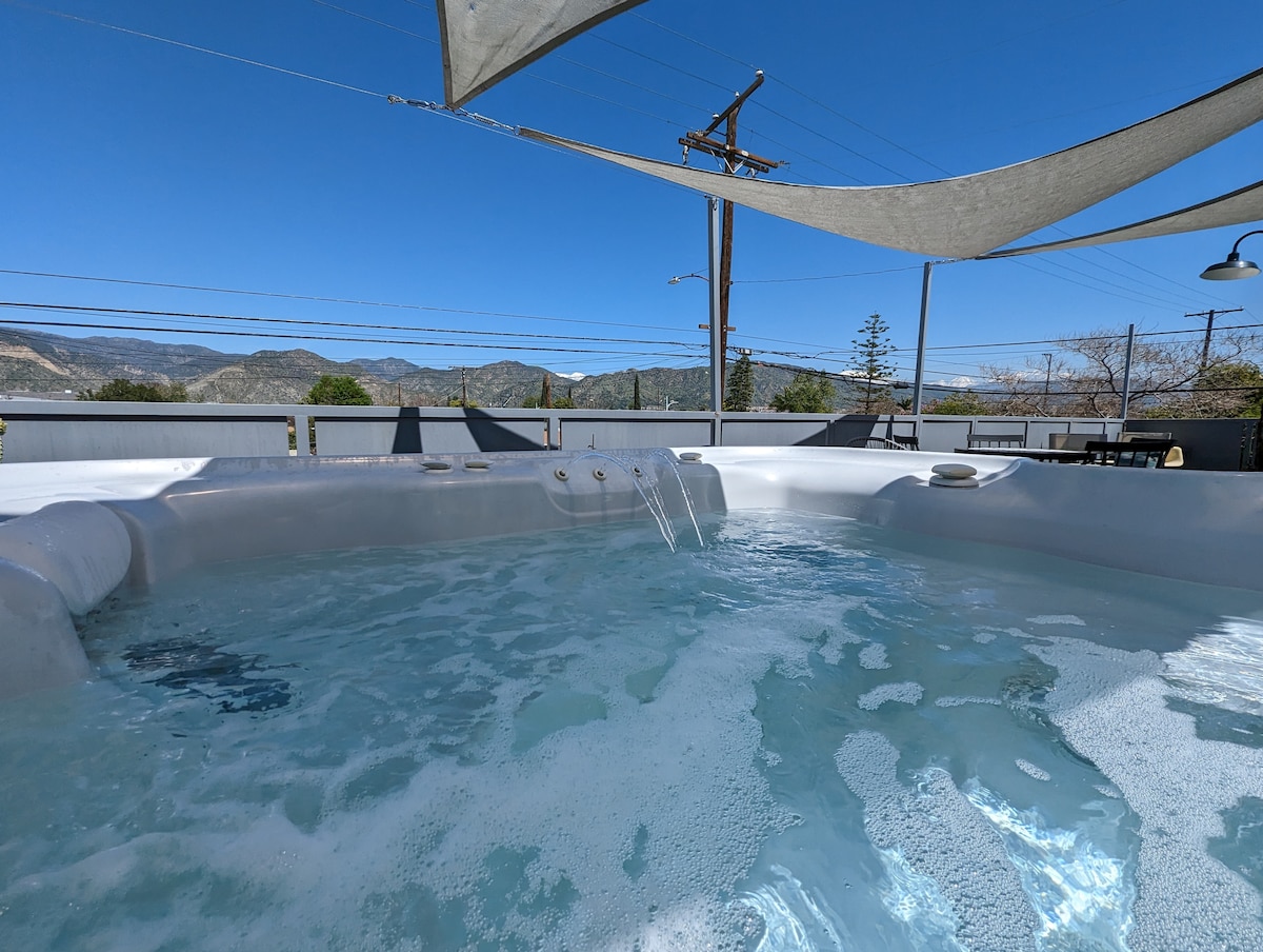 A hot tub is filled with bubbling water, surrounded by a spacious deck and set against a backdrop of mountains. Comfortable lounge chairs are positioned nearby, and a shade sail provides protection from the sun.