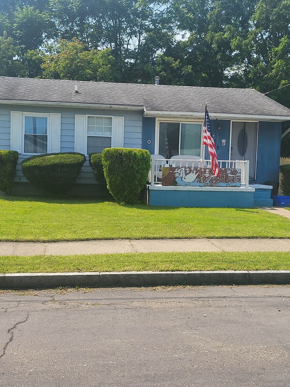 A single-story home is presented, featuring a light blue exterior and a covered porch adorned with a white railing. An American flag is displayed prominently. Neatly trimmed hedges line the front yard, while a well-maintained lawn offers a vibrant green appearance.