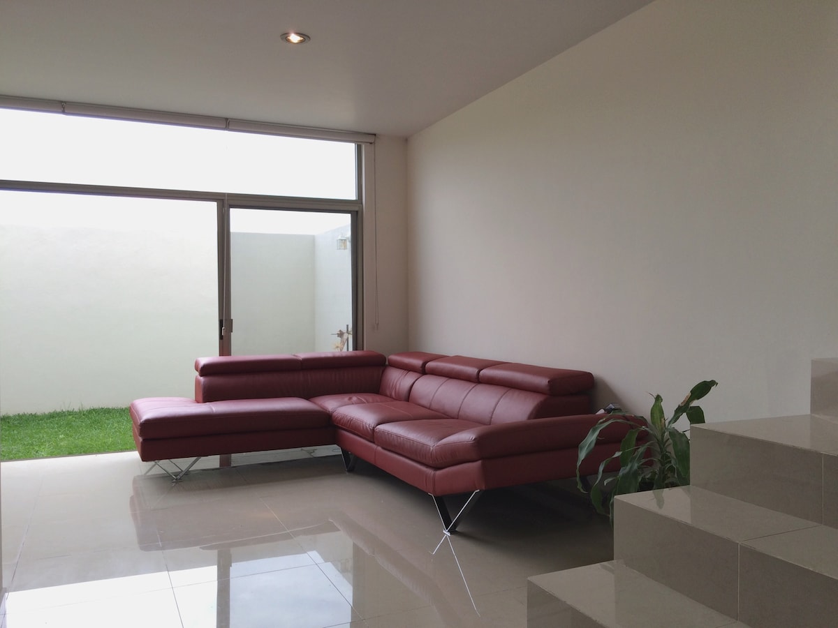 A modern living area features a red sectional sofa positioned near large glass windows, allowing natural light to fill the space. Green grass is visible outside, creating a connection to the outdoor environment. Smooth tile flooring complements the contemporary aesthetic.