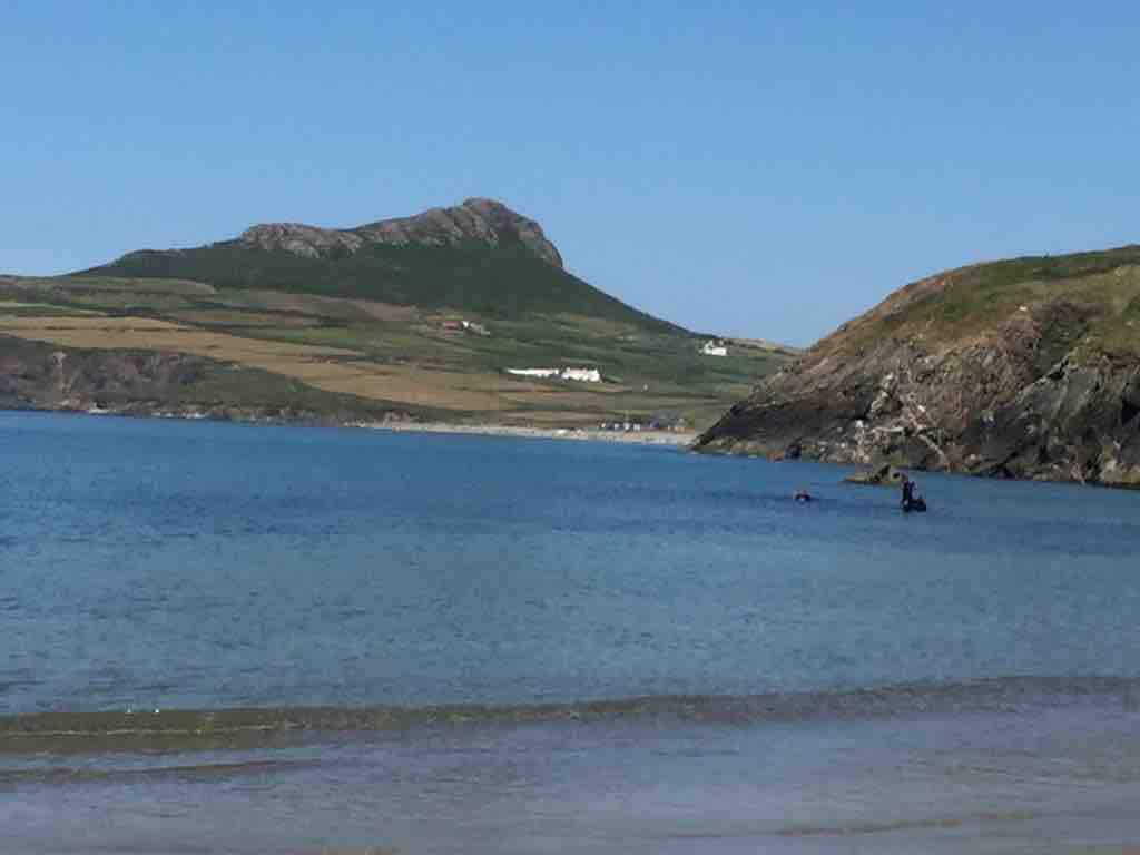 The image captures a serene beach scene featuring calm waters gently lapping at the shore. A distinctive rocky hill rises in the background, framed by lush green fields, while a few distant white structures are partially visible on the hillside.