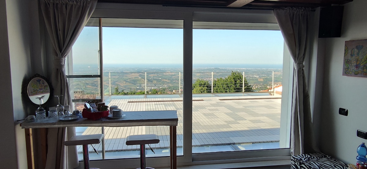 A large window offers panoramic views of the surrounding landscape, showcasing rolling hills and the sea in the distance. A small table with two chairs and a tray of refreshments is positioned nearby, complementing the bright and airy atmosphere of the room.