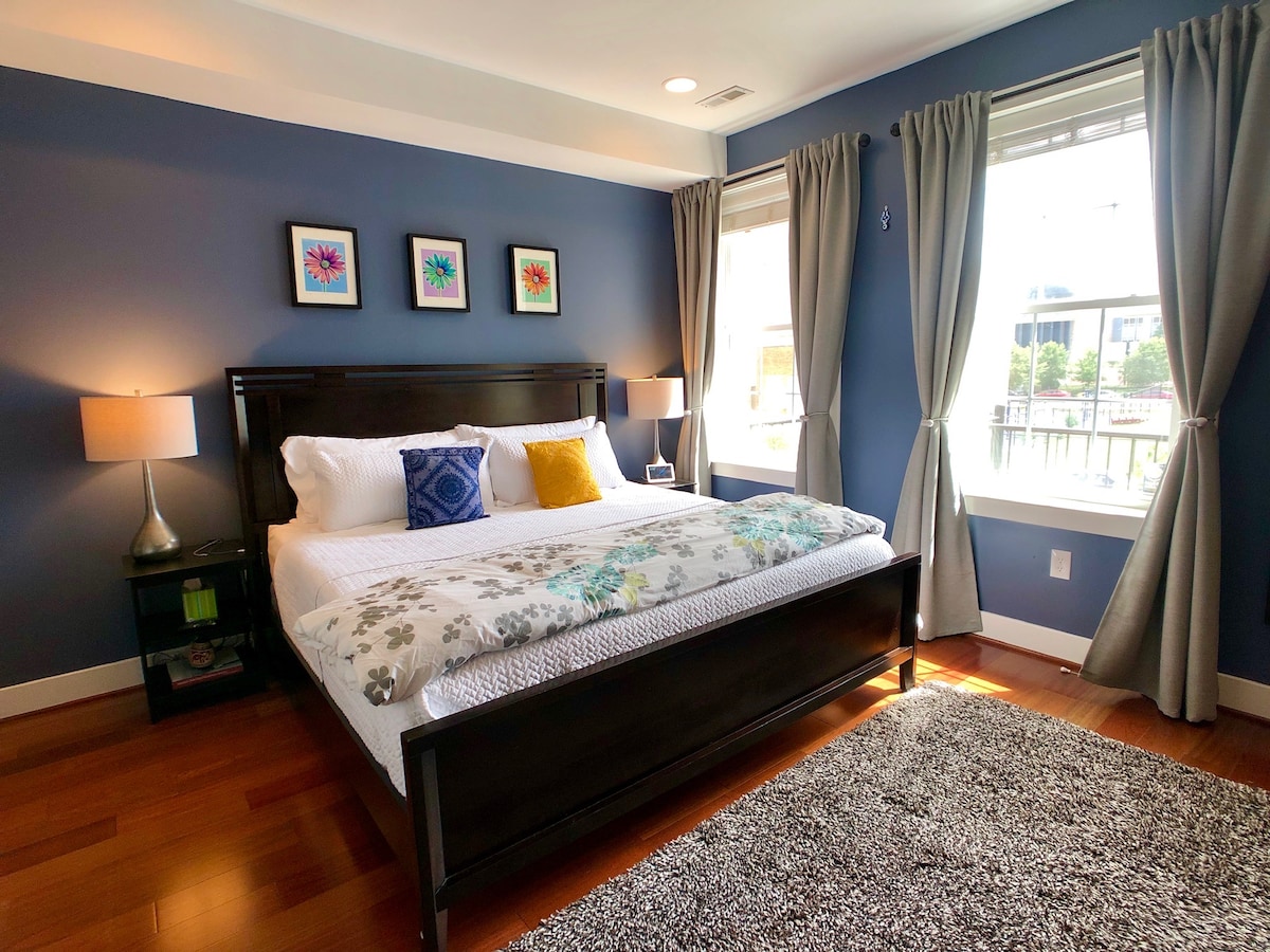 A spacious bedroom features a comfortable king-sized bed, dressed in soft linens. Framed floral artwork adorns the blue walls, complemented by large windows with light-filtering curtains. Two bedside lamps provide soft illumination, while a plush area rug adds warmth to the hardwood floor.