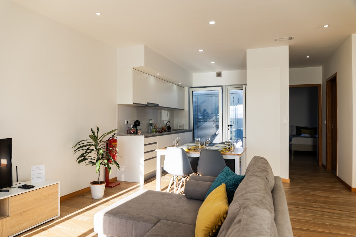 A modern living area showcases a comfortable sofa adorned with colorful cushions. A dining table is set for four, positioned near large glass doors that provide access to an outdoor space. Natural light brightens the open layout, emphasizing the sleek kitchen in the background.