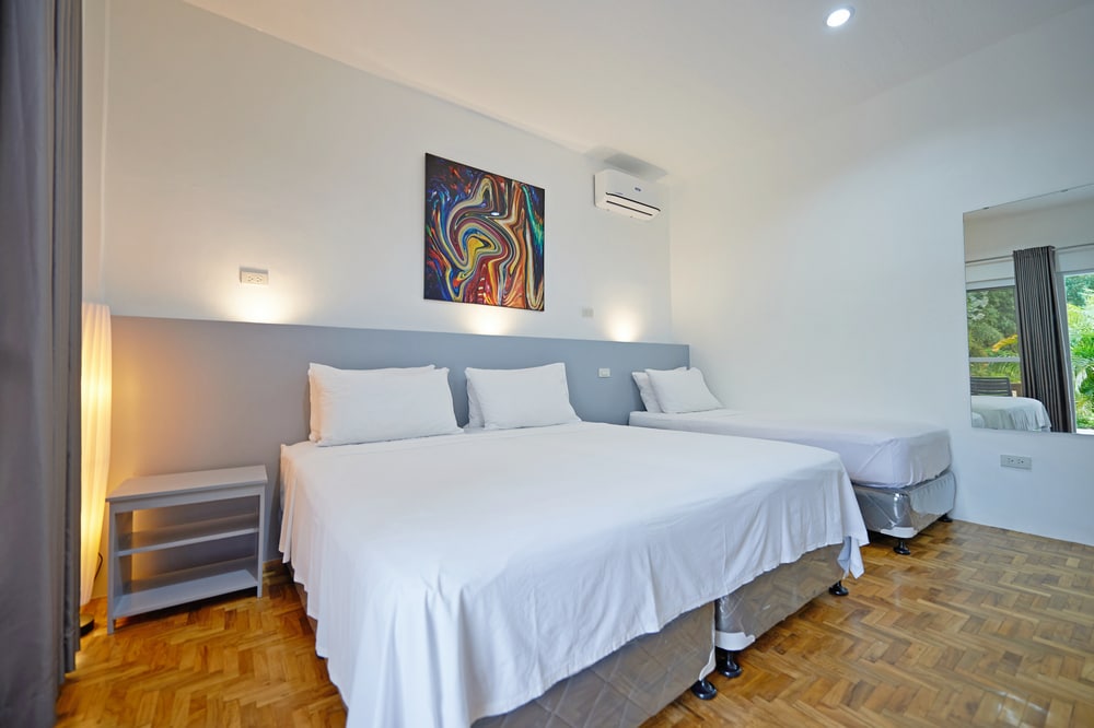 A spacious room is presented with two large beds, each dressed in crisp white linens. A vibrant piece of wall art adds color to the neutral tones of the room. Natural light streams in from a window, complemented by soft lighting from bedside lamps.