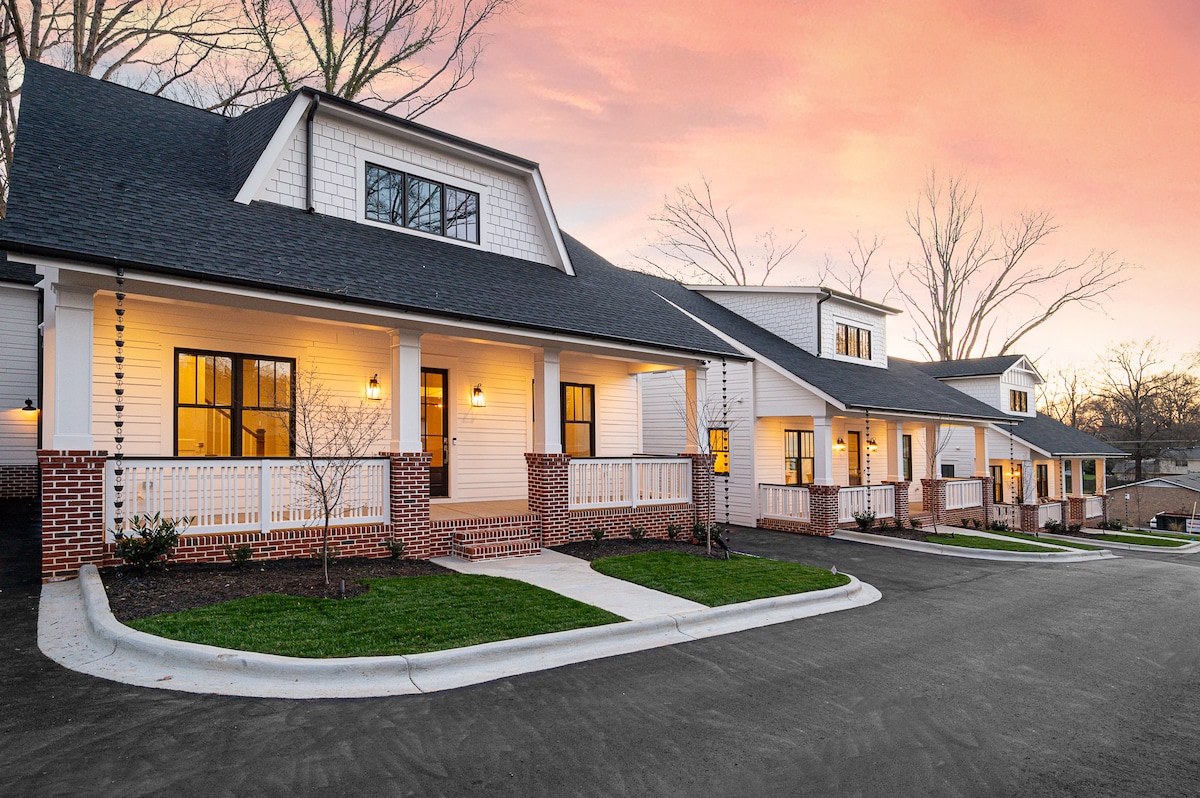 A row of charming cottages is set against a colorful sunset sky. Each unit features a welcoming porch with inviting lighting, surrounded by well-maintained landscaping. The smooth concrete drive curves gently alongside the cottages, providing a sense of accessibility and neighborhood warmth.