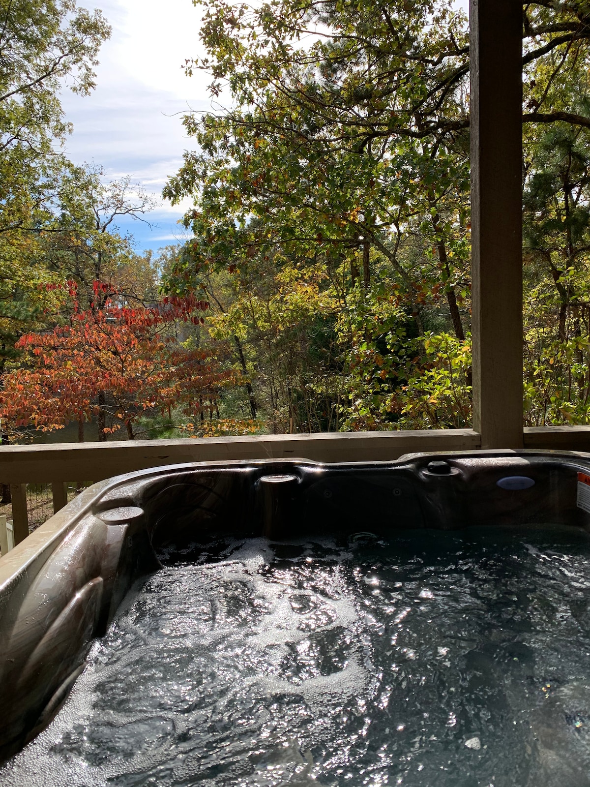 A hot tub is situated on a wooden deck, with bubbles creating ripples on the surface of the water. Trees in vibrant autumn colours surround the area, offering a serene view of the natural landscape beyond.