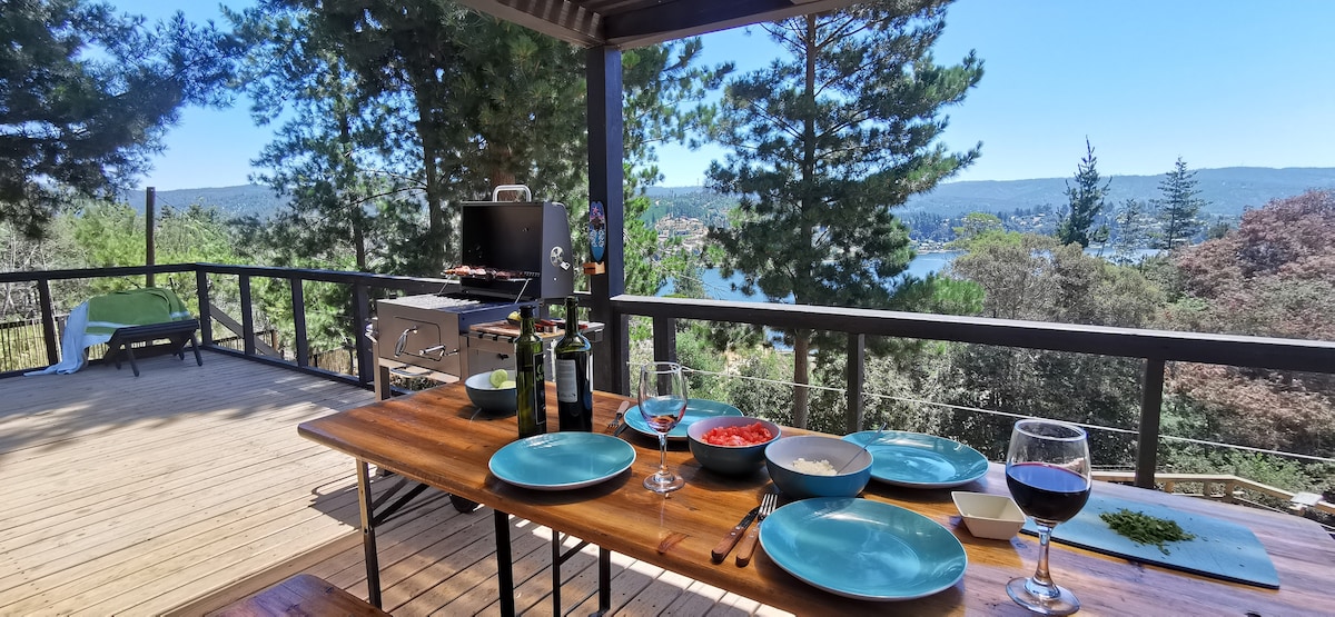 A spacious deck features a dining table set for several guests, complete with plates and wine glasses. A grill is positioned near the edge, offering views of the lake and surrounding forest, creating an inviting outdoor dining space.