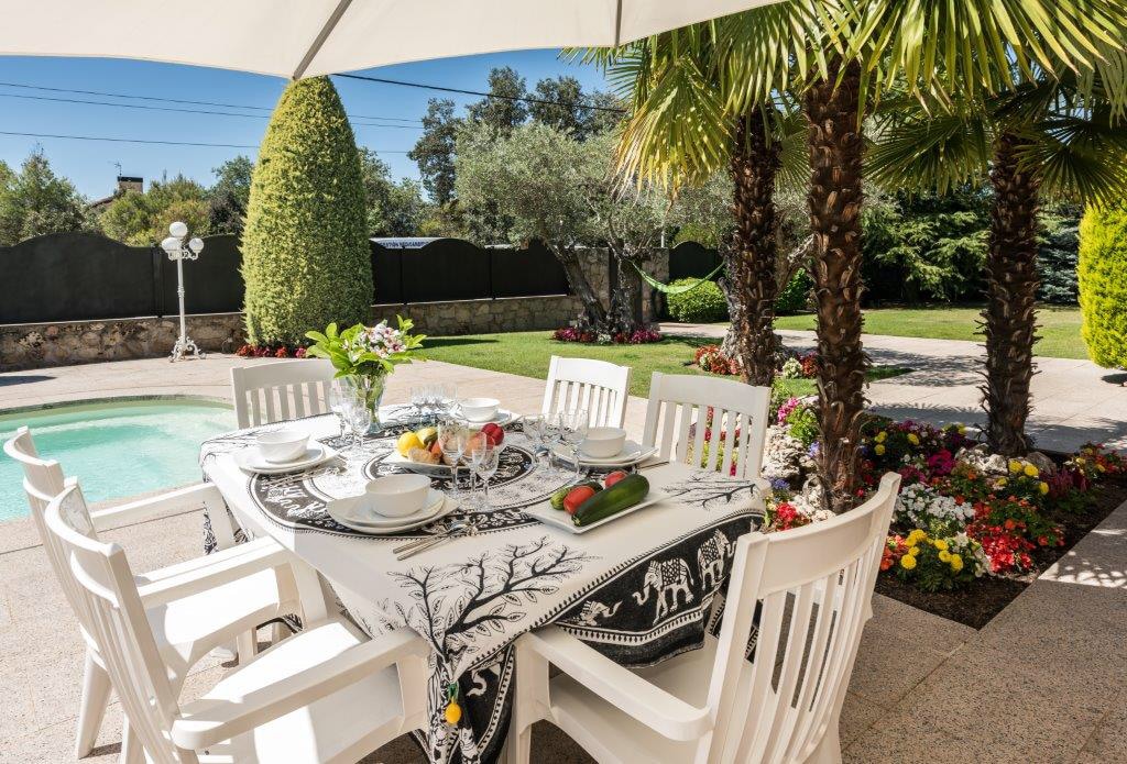 An outdoor dining area features a table set for entertaining, surrounded by eight white chairs. The setting is complemented by a beautifully manicured garden with colorful flowers and palm trees, with a swimming pool visible in the background under a sunny sky.