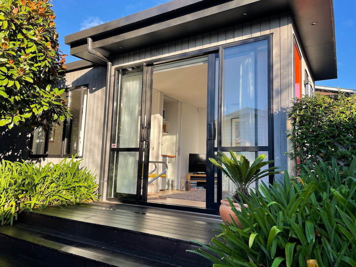 A modern apartment exterior is captured, featuring large black-framed glass doors that open to a bright living area. Lush greenery surrounds the entrance, and a wooden deck leads to the doorway, inviting a seamless connection between indoor and outdoor spaces.