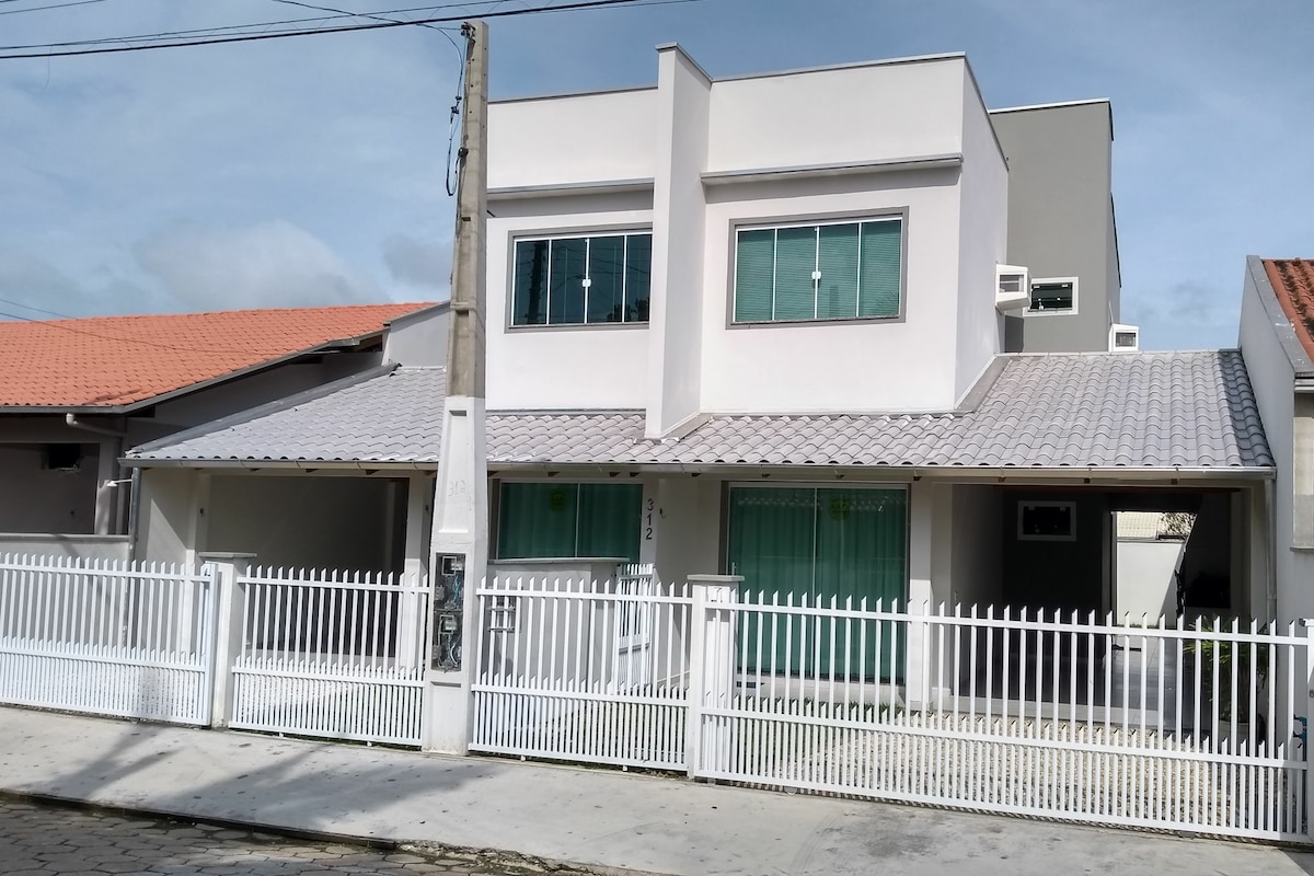 The exterior of the recently constructed house is captured, showcasing a two-story design with large windows. A white picket fence delineates the property, complemented by a paved walkway and decorative roof tiles. Neighboring structures and a clear sky are visible in the background.