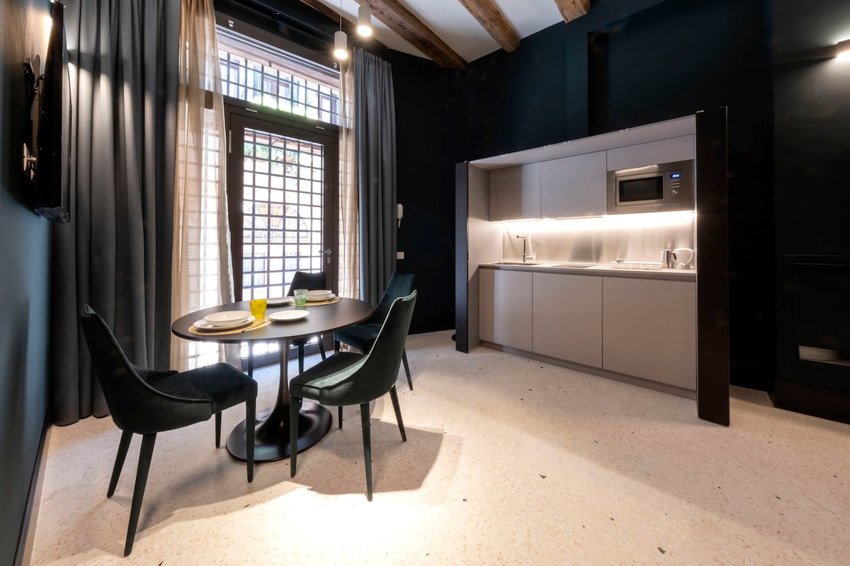 A contemporary dining area is complemented by a round table surrounded by four dark green chairs. The well-equipped kitchenette features a microwave and countertop lighting, while large windows with light curtains offer natural illumination. Exposed wooden beams enhance the character of the space.