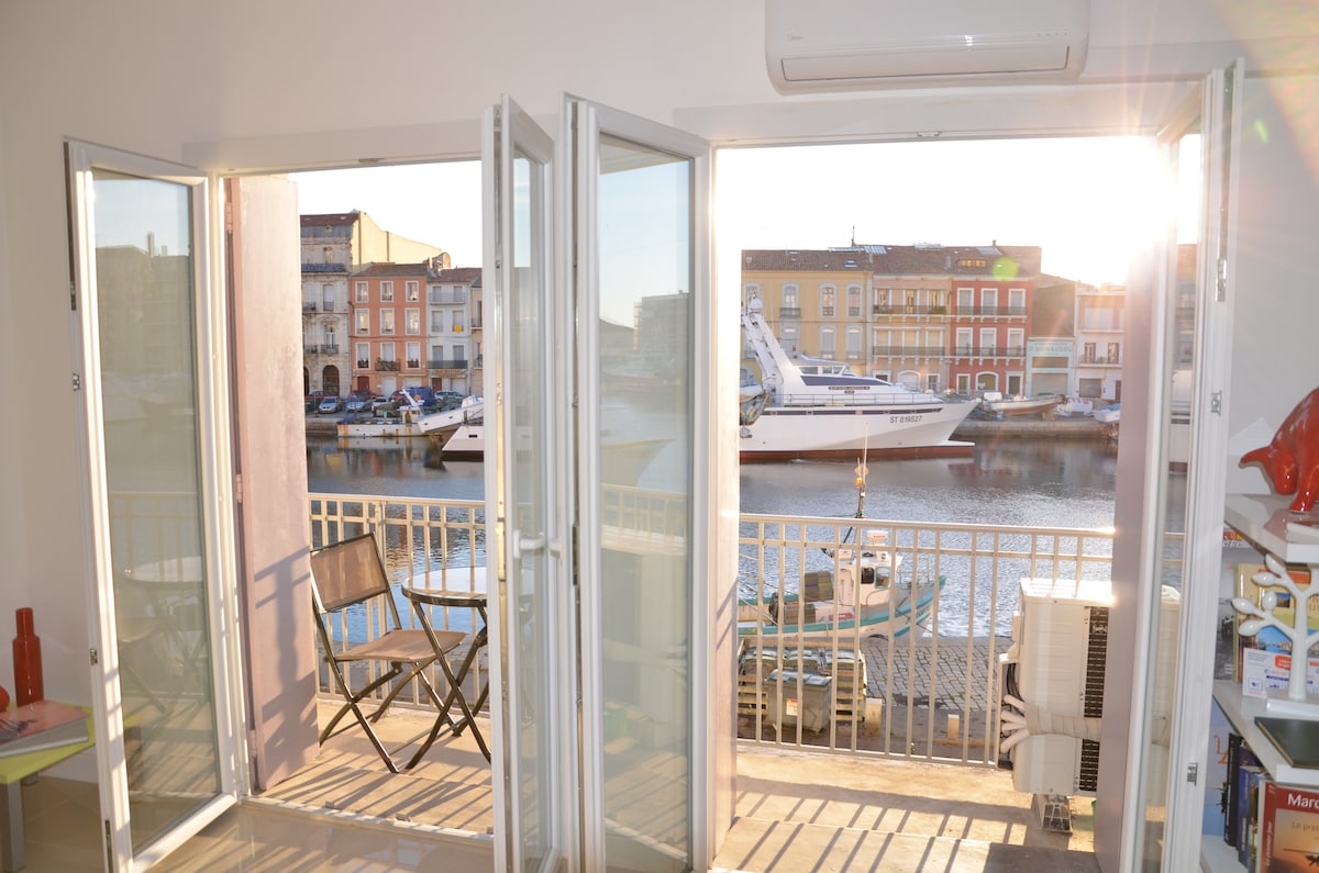 Broad glass doors open to a balcony overlooking the canal. A single chair is positioned on the balcony, while the sunlight reflects off the water, creating a warm ambiance. The vibrant colors of adjacent buildings and a moored boat are visible in the background.