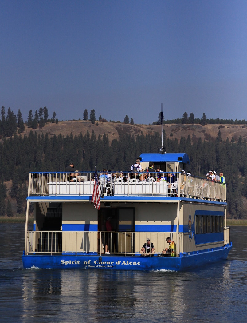 A sightseeing boat named 'Spirit of Coeur d'Alene' is shown cruising on calm waters, surrounded by green hills. The upper deck accommodates several guests enjoying the panoramic views, while a flag flutters on the top. The boat features a two-level structure with an open-air design.