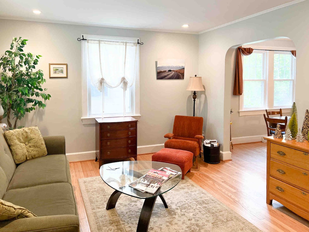 A comfortable living area features a light green couch and a red armchair, with a glass coffee table at the center. Natural light filters through a sheer-curtained window, illuminating a side table and a wooden dresser. A decorative plant adds a touch of green to the space.
