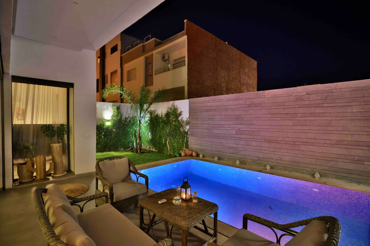 An outdoor seating area is visible next to a swimming pool, illuminated with soft blue lighting. Potted plants add greenery to the space, while a low table with lanterns and refreshments creates a welcoming atmosphere for relaxation.
