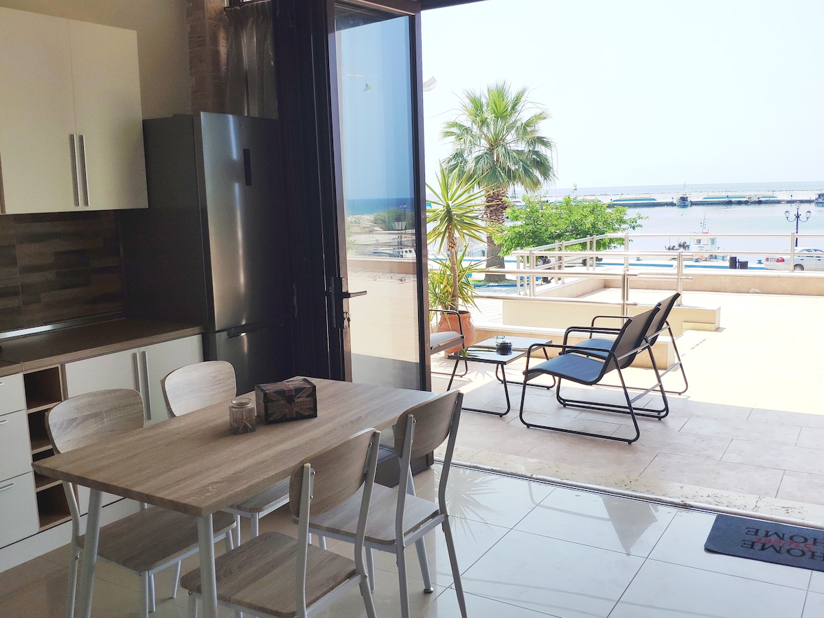 A dining area features a wooden table with white chairs, positioned near large glass doors that open to a patio. Outside, a sun lounger is visible, with palm trees and the harbor in the background, providing a bright and airy atmosphere.