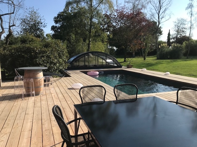 A wooden deck extends around a heated swimming pool, featuring an attached retractable cover. The scene includes a black dining table surrounded by chairs, with a barrel nearby. Lush greenery and trees provide a natural backdrop, creating an inviting outdoor space.