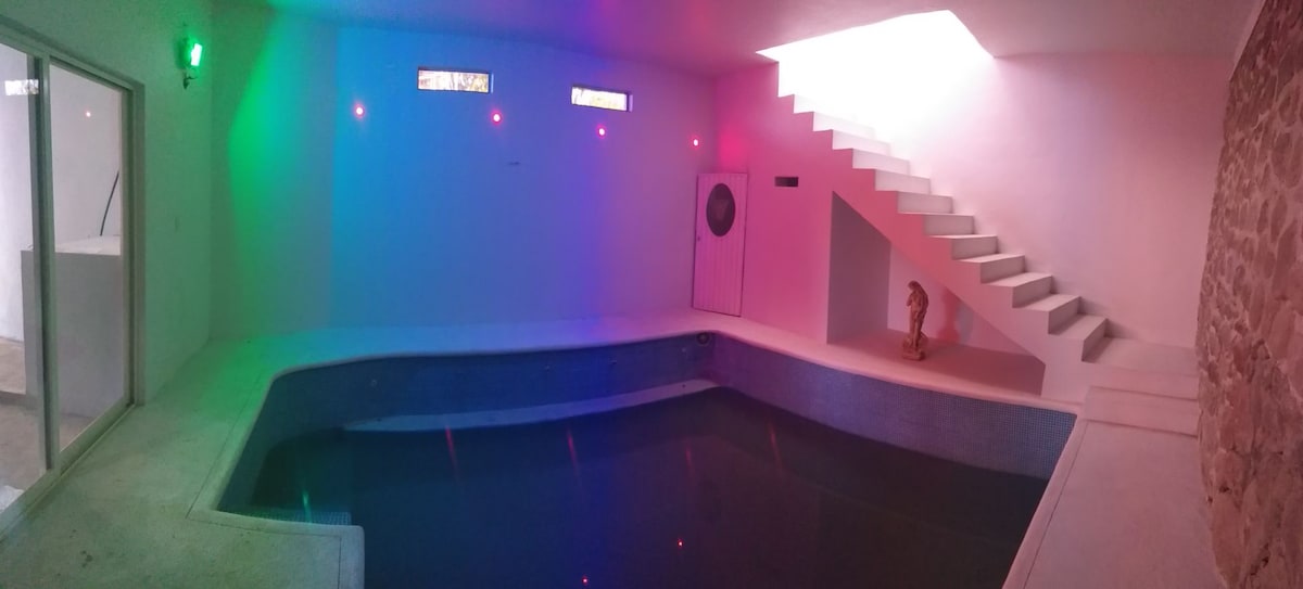 An indoor heated pool is showcased with a sleek, modern design. Soft, colorful lighting gently illuminates the water, while natural light filters through a skylight above. A staircase is visible on one side, leading to the upper level of the home.