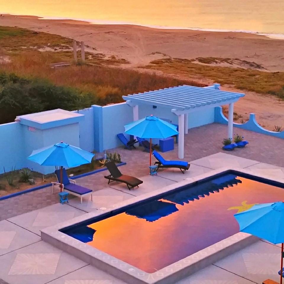 The scene features a tranquil private pool surrounded by vibrant blue umbrellas and lounge chairs, reflecting the colorful hues of a sunset. A sandy beach is visible in the background, blending into a serene coastal environment.
