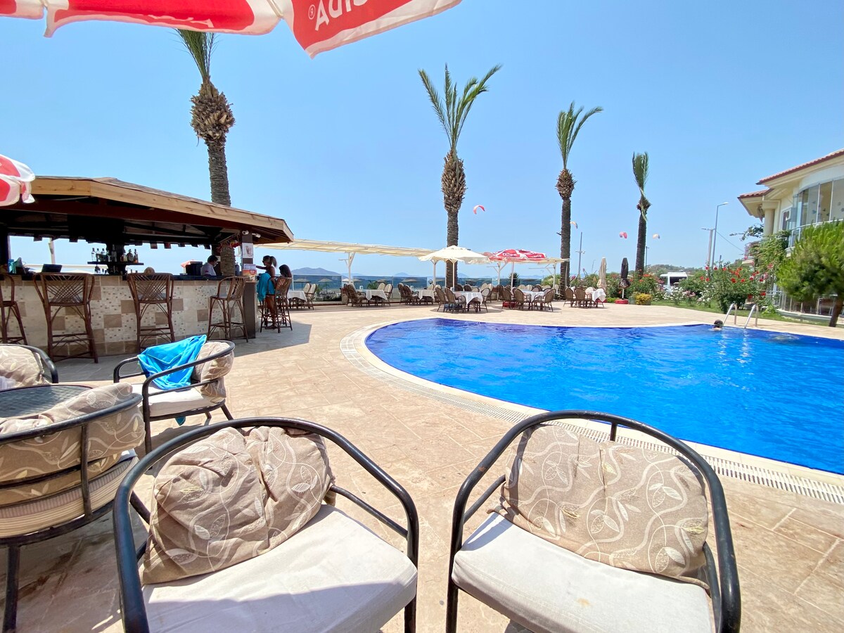 A relaxing poolside area is presented, featuring two comfortable chairs with soft cushions. A vibrant blue pool reflects the clear sky, while palm trees sway gently in the background. An outdoor bar is visible, creating a leisurely atmosphere for guests.