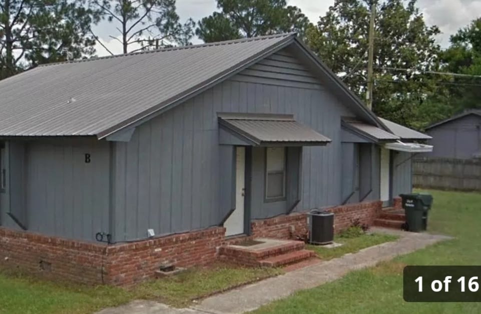 A gray single-story building is shown, featuring two separate entries with doorways surrounded by white trim. The structure is set against a backdrop of green trees, with a brick foundation and steps leading to each entrance. A trash bin is visible beside the building.