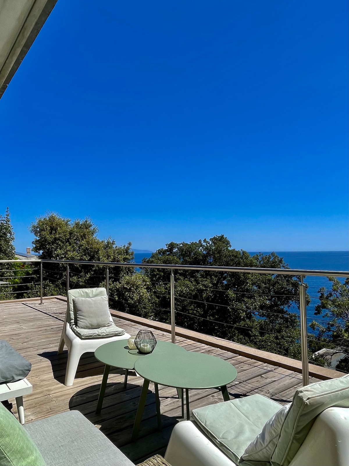 A spacious terrace with a panoramic view of the sea features two white lounge chairs and a green table. A soft blue sky and lush greenery create a serene backdrop, inviting relaxation in the coastal environment.